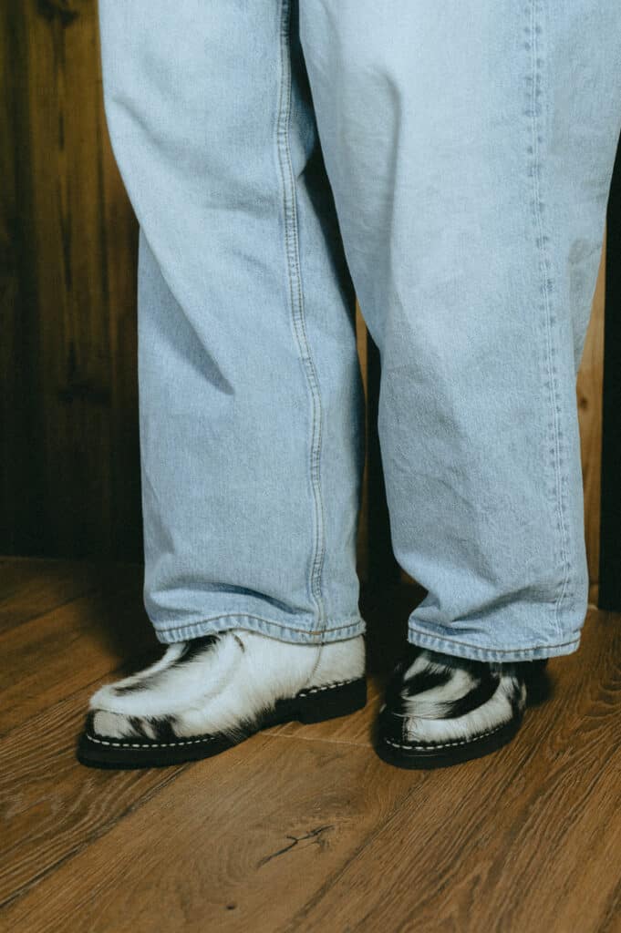 Femme portant des Paraboot Michael poils de vache et un jean large dans un intérieur type chalet avec cheminée.