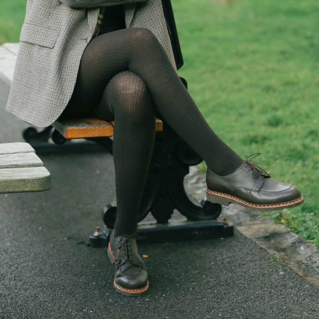 Femme assise sur un banc de parc portant des chaussures Paraboot Chamade en cuir lisse moka, associées à des collants noirs et une veste à carreaux.