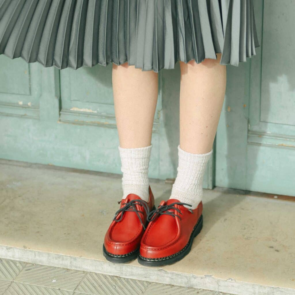 Femme debout portant des chaussures Paraboot Michael en cuir lisse rouge, une jupe plissée verte et des chaussettes écrues.