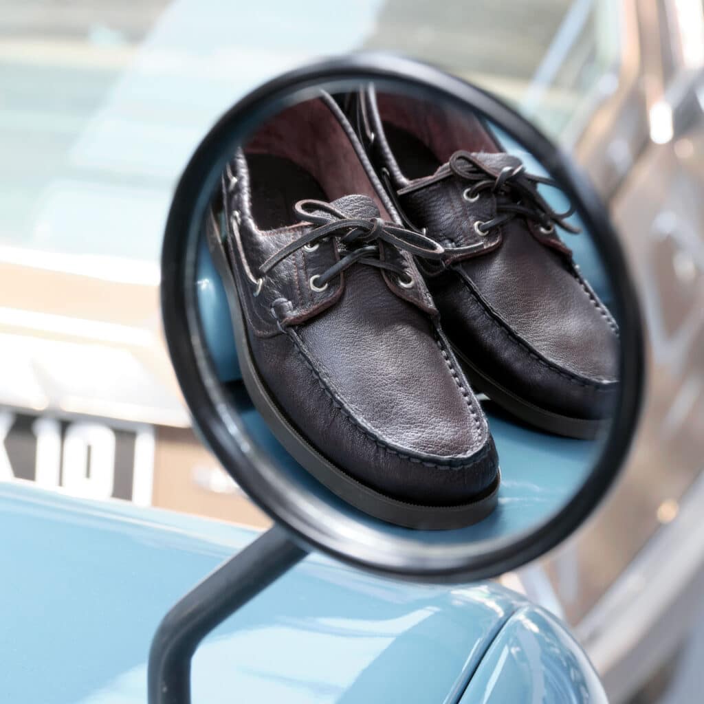 Chaussures bateau Paraboot en cuir marron foncé reflétées dans le rétroviseur et posées sur l'aile d'un 4x4 ancien.