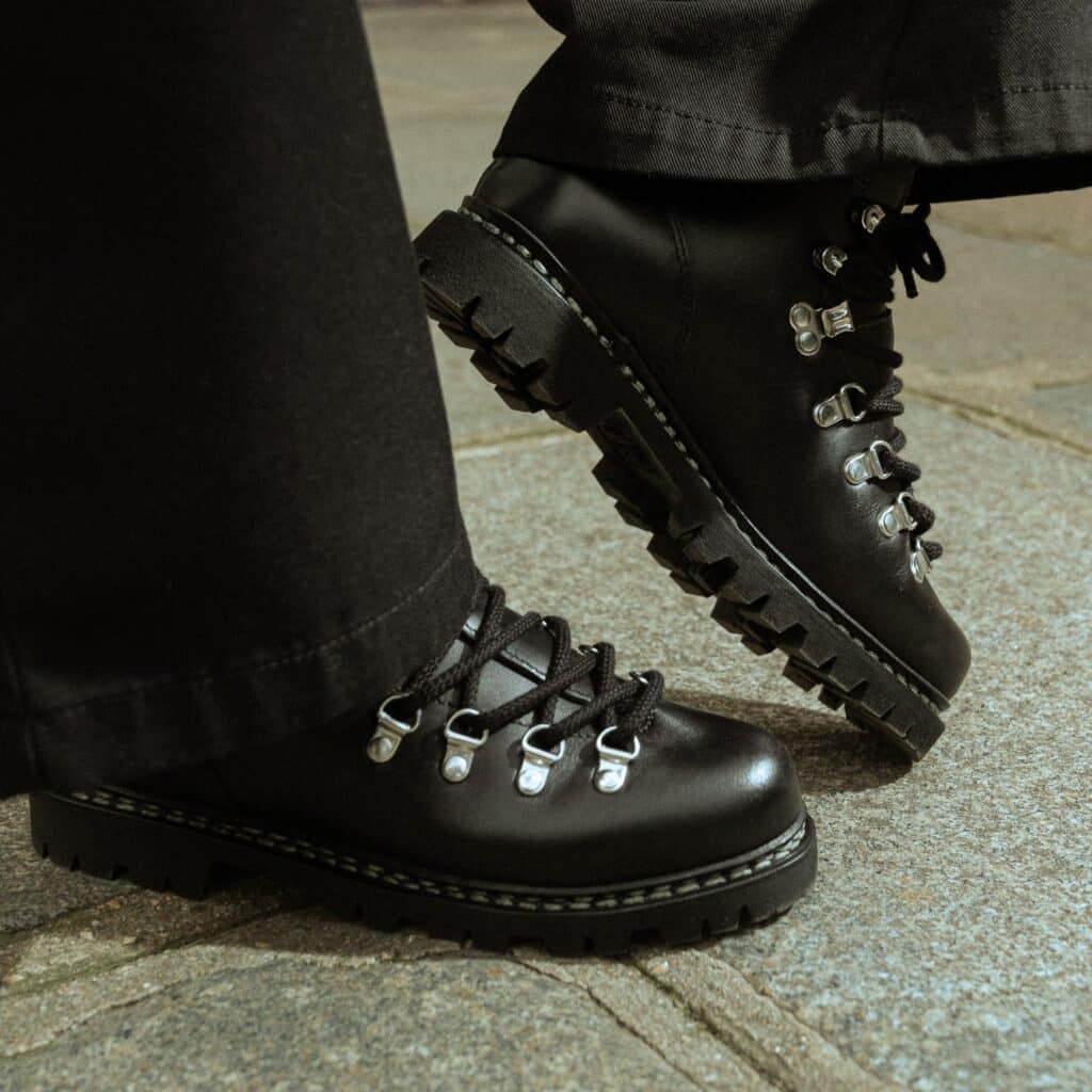 Gros plan de bottines de ville femme Paraboot Avoriaz en cuir lisse noir avec semelle commando et laçage montagne, portées sur pavés avec un pantalon noir.
