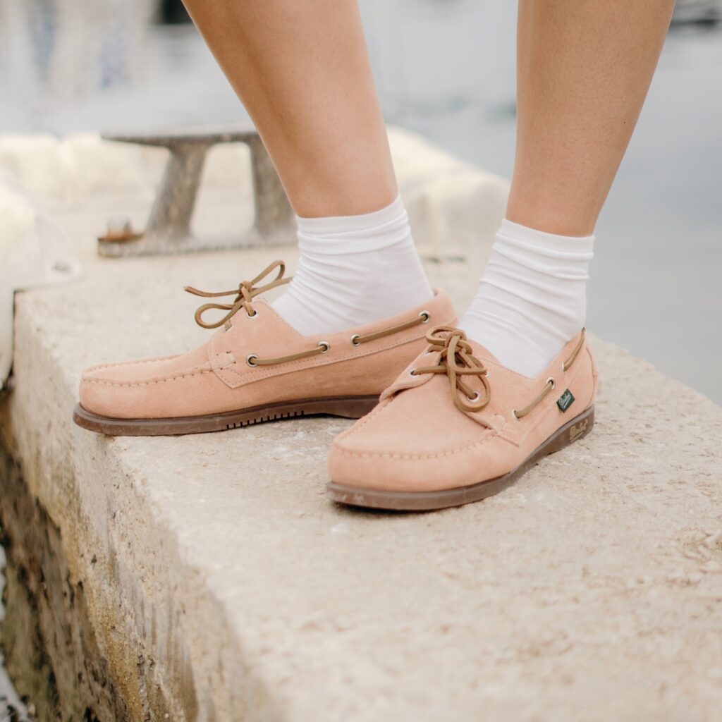 Gros plan sur les pieds d'une personne portant des chaussures bateau Barth de Paraboot en velours nude avec des chaussettes blanches, debout sur un quai en pierre au bord de l'eau.