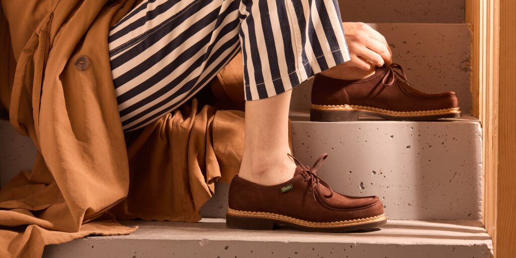 Mirabelle en cuir nubuck Taurillon portée par une femme assise. L'association avec un trench coat en velours et un pantalon blanc à rayure marron souligne parfaitement le confort et l'élégance couplé au savoir-faire artisanal de Paraboot.