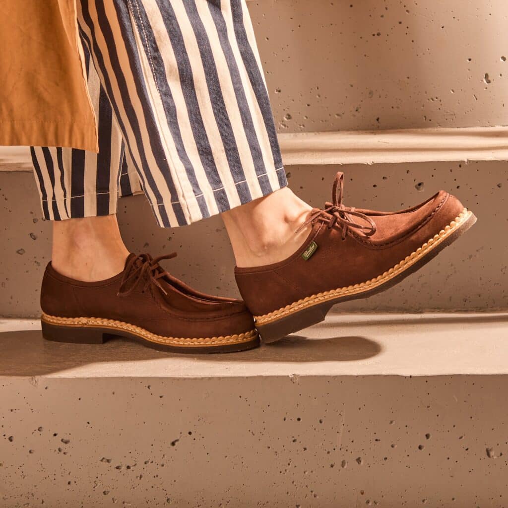 Zoom sur les chaussures à lacets Mirabelle de Paraboot en nubuck taurillon chocolat, portées sur un escalier en béton avec un pantalon à rayures.