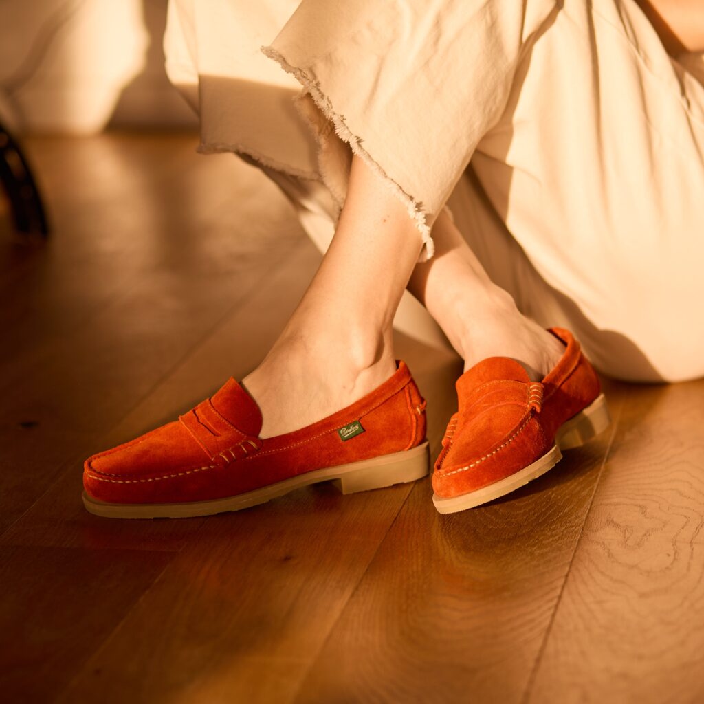Gros plan sur les pieds d'une femme assise sur un parquet en bois, portant des mocassins Coraux de Paraboot en velours koi de couleur orange avec une étiquette verte.