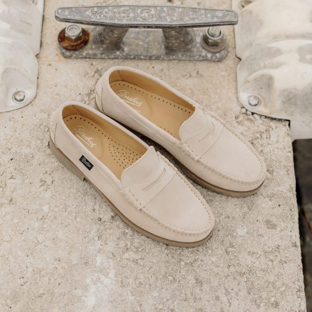 Vue de dessus d'une paire de mocassins Coraux de Paraboot en velours crème, posée sur un quai en pierre à côté d'un taquet d'amarrage métallique.