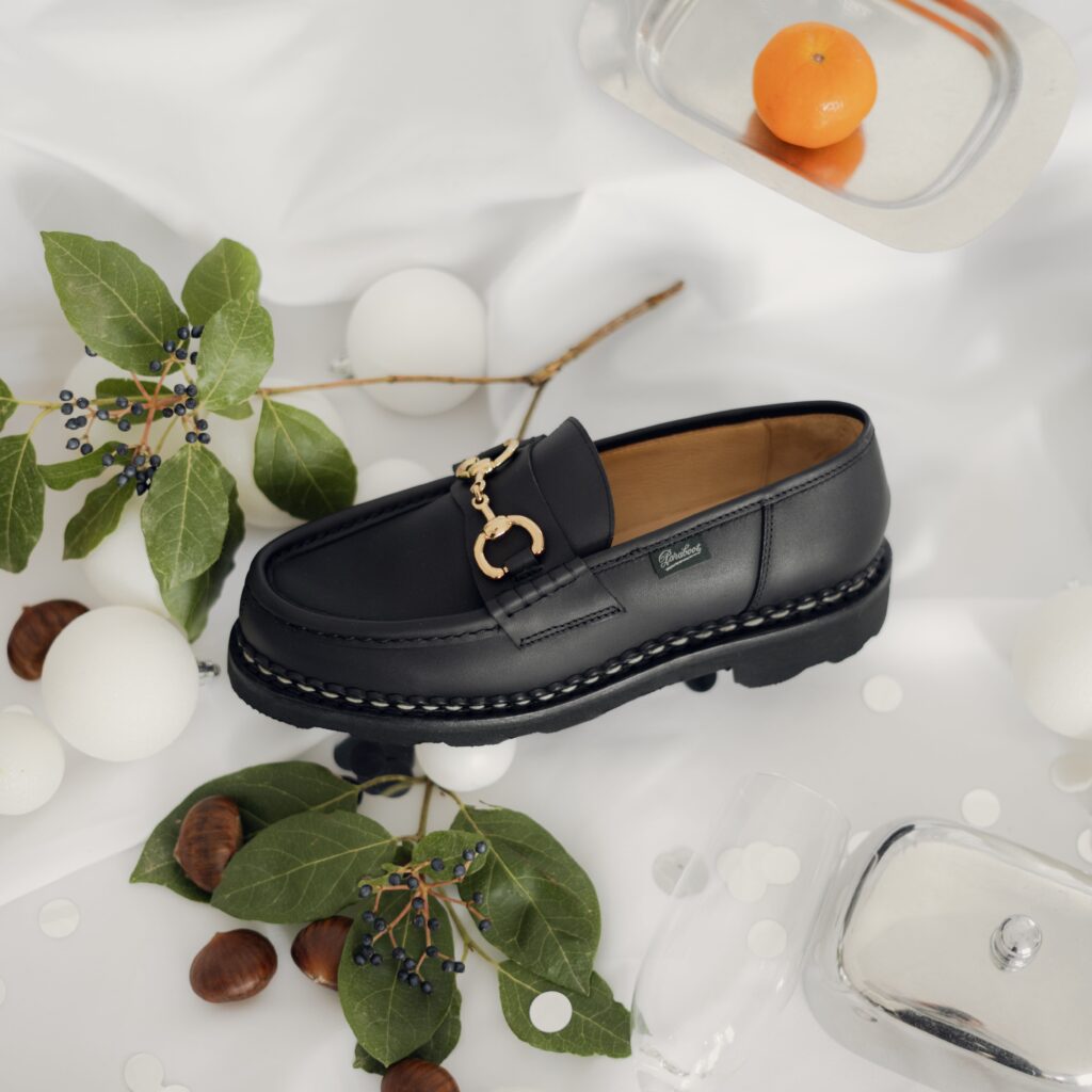 Orsayti de Paraboot en cuir lisse noir avec mors doré, présentés sur un drapé blanc à côté d'une boîte Paraboot et d'une bougie.