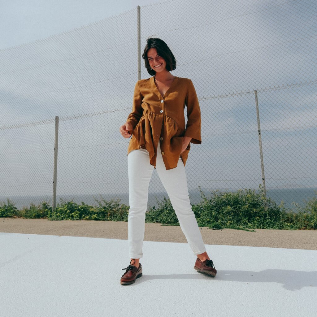 Femme souriante en extérieur portant des chaussures bateau Barth de Paraboot en cuir marron America, pantalon blanc et veste moutarde.
