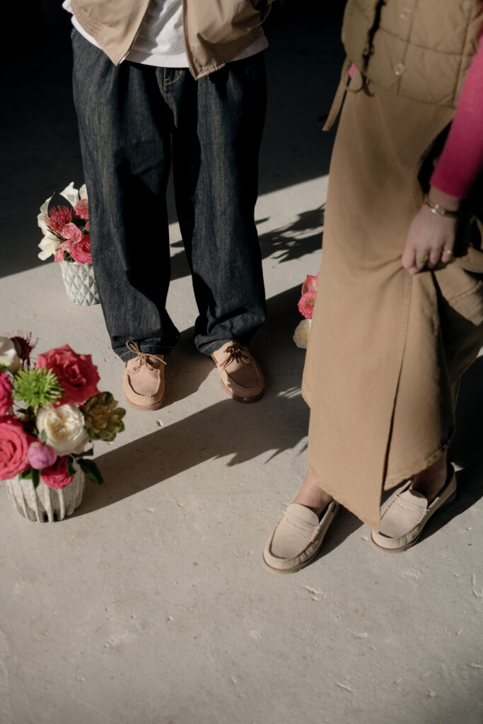 Chaussures été femme : vue de dessus de deux personnes portant des mocassins Coraux en velours crème et la chaussure bateau barth en velours nude Paraboot entre des pots de fleurs colorées.
