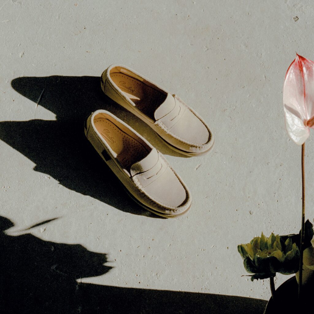 Paire de mocassins Coraux de Paraboot en velours crème posée sur un sol en béton clair à côté d'une fleur d'anthurium.