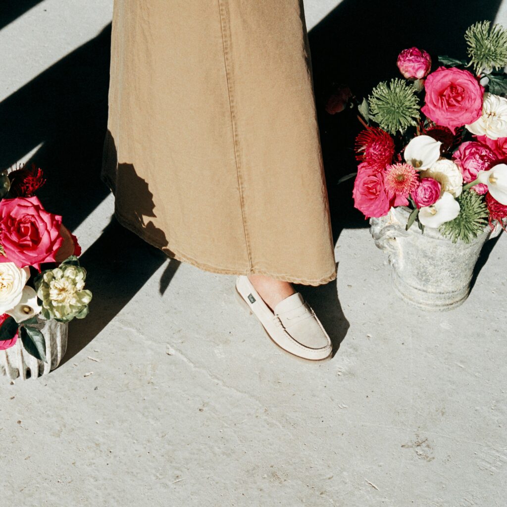 Vue de dessus d'une personne portant un mocassin Coraux de Paraboot en velours crème avec une jupe longue beige, marchant entre des pots de fleurs colorées.