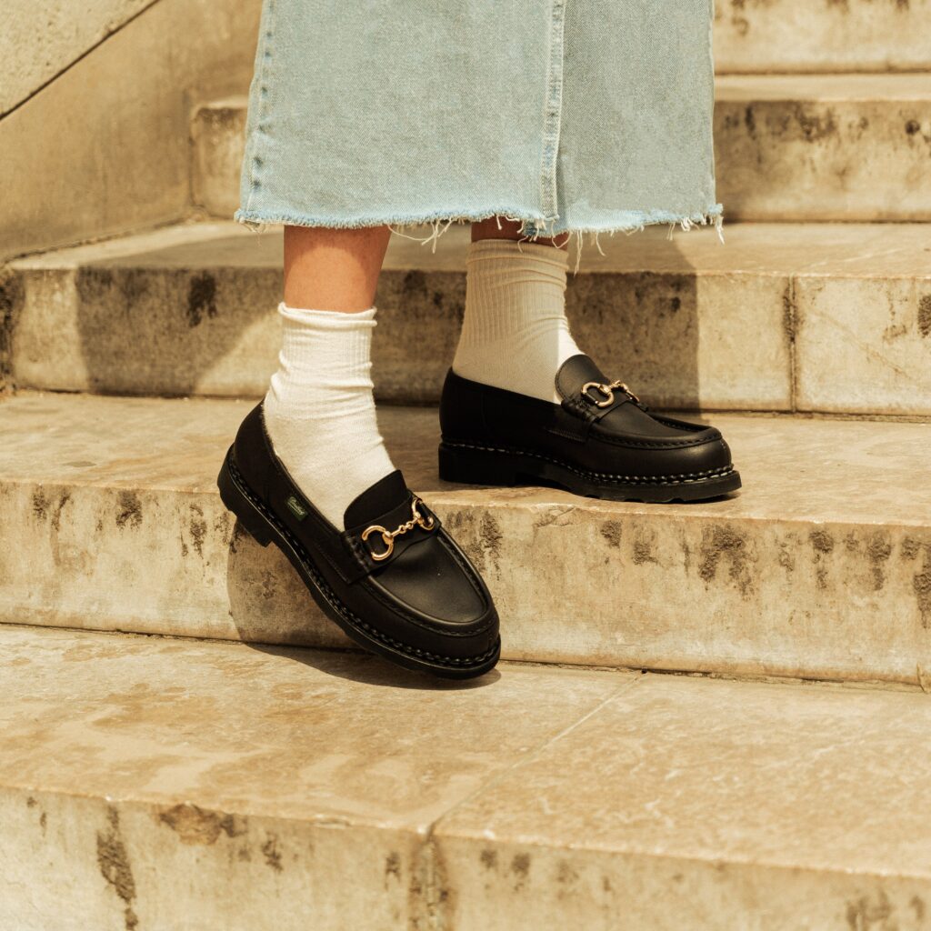Gros plan sur les pieds d'une femme portant des mocassins Orsayti de Paraboot en cuir noir avec mors doré, des chaussettes blanches et une jupe en jean, descendant un escalier en pierre.