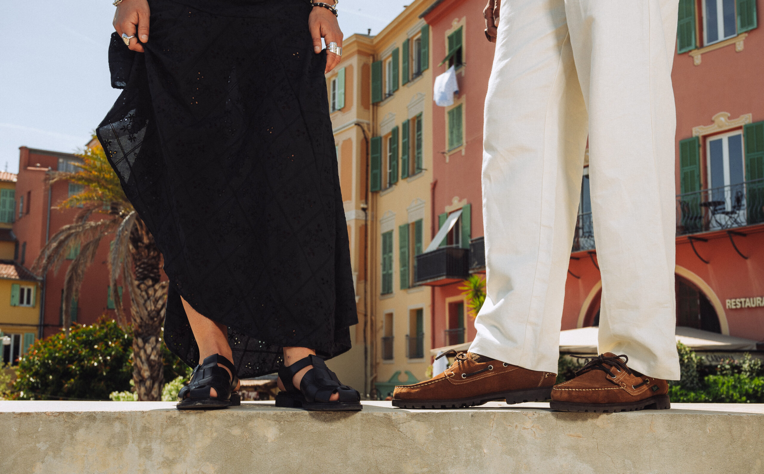 Sandales en cuir noir Ibéris pour femme et chaussures bateau Malo en velours cognac pour homme Paraboot, portées en extérieur devant des façades colorées du Sud.