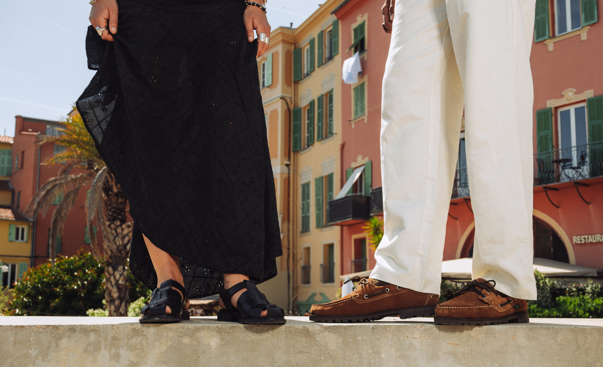 Paraboot women's black leather Ibéris sandals and men's cognac velvet Malo boat shoes, worn outdoors in front of colorful facades in the South.