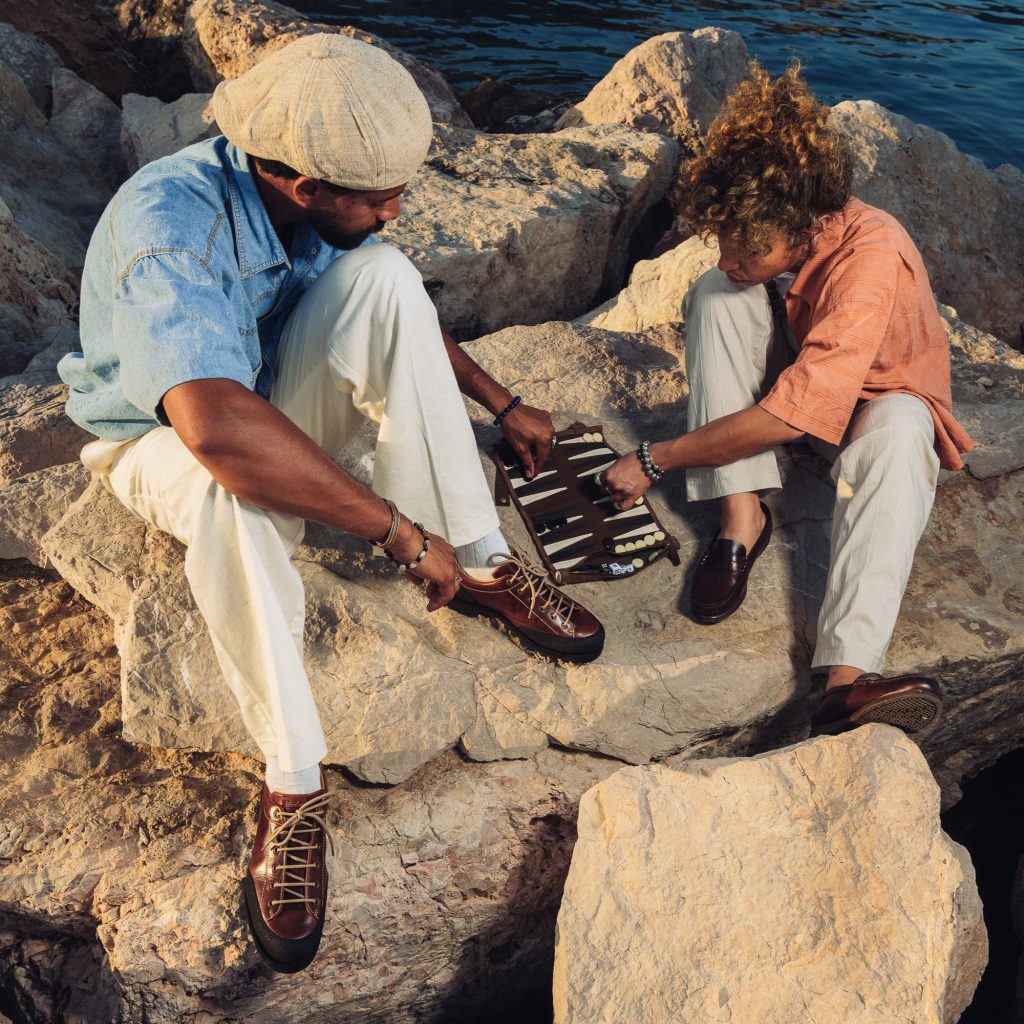 un couple jouant au backgammon sur les rochers en bord de mer, l'homme est chaussé avec le derby Montana et la femme porte le modèle Vignony lisse café Paraboot.