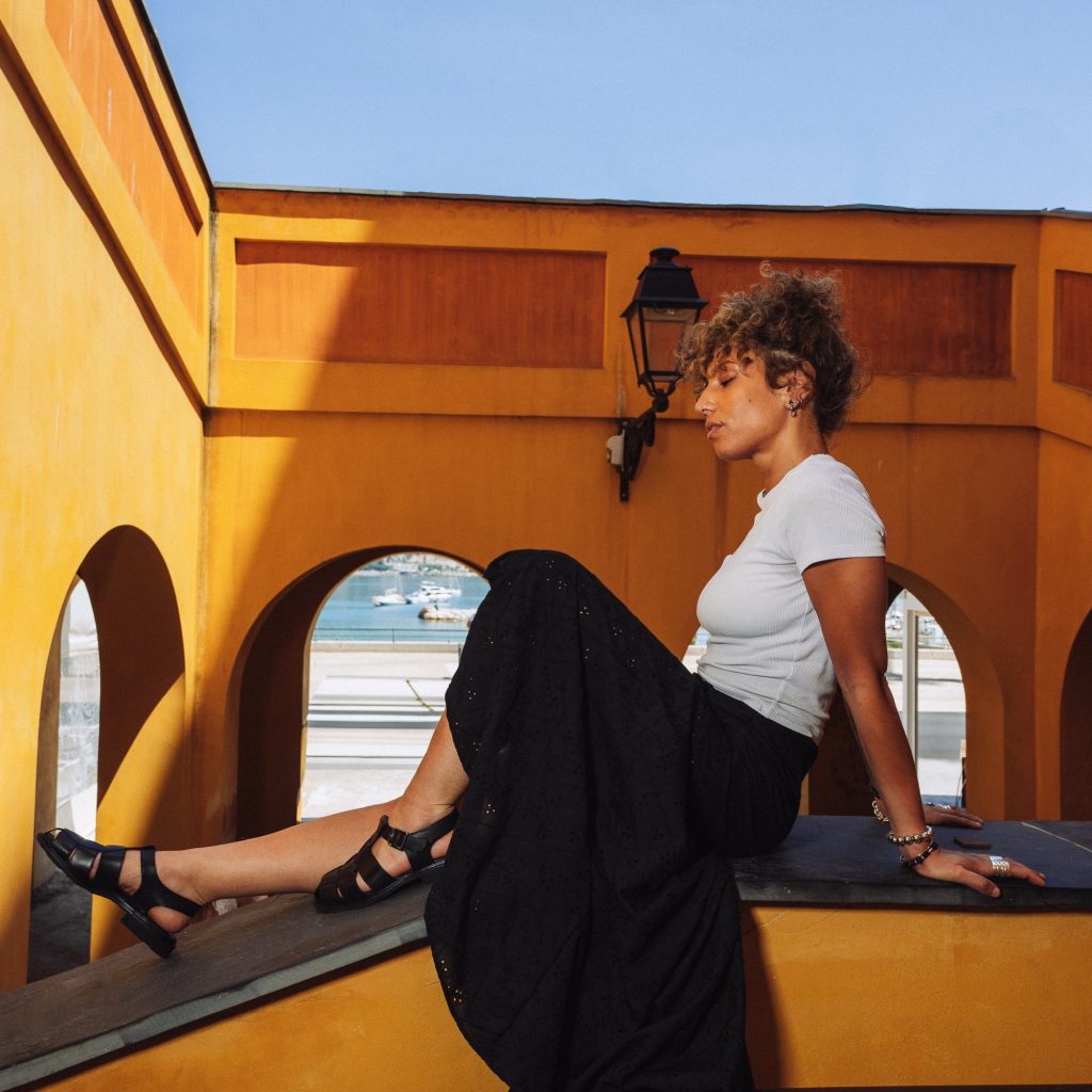 Femme assise sous les arcades ocre de Menton portant des sandales tressées Iberis noires Paraboot. L'élégance intemporelle de la sandale Iberis, sous le soleil de la Riviera.