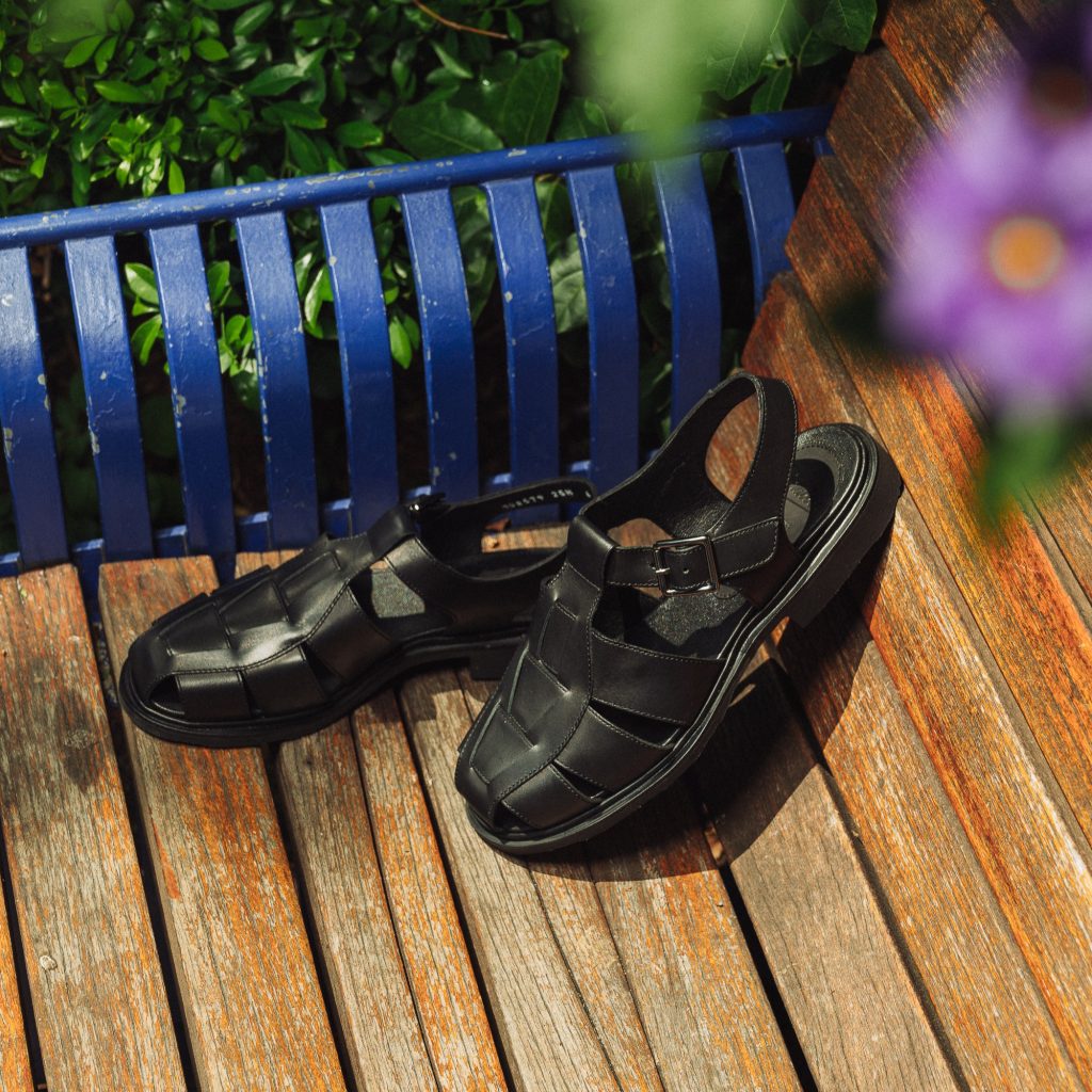 Sandales en cuir noir tressé Iberis Paraboot posées sur un banc en bois au soleil. Le modèle Iberis : l'équilibre parfait entre aération estivale et maintien du pied.
