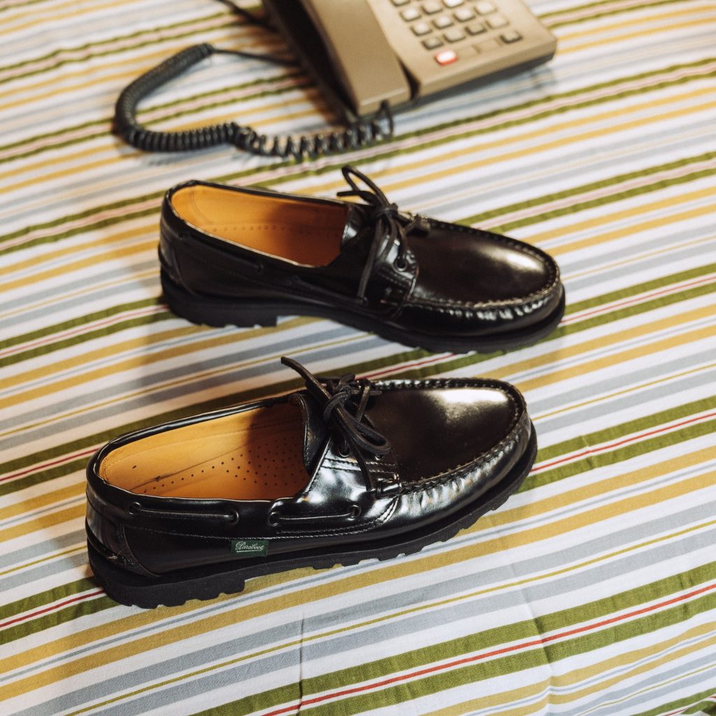 Chaussures bateau Malo en cuir brillant noir Paraboot sur un lit aux draps rayés. Brillance et robustesse : l'incontournable modèle Malo.