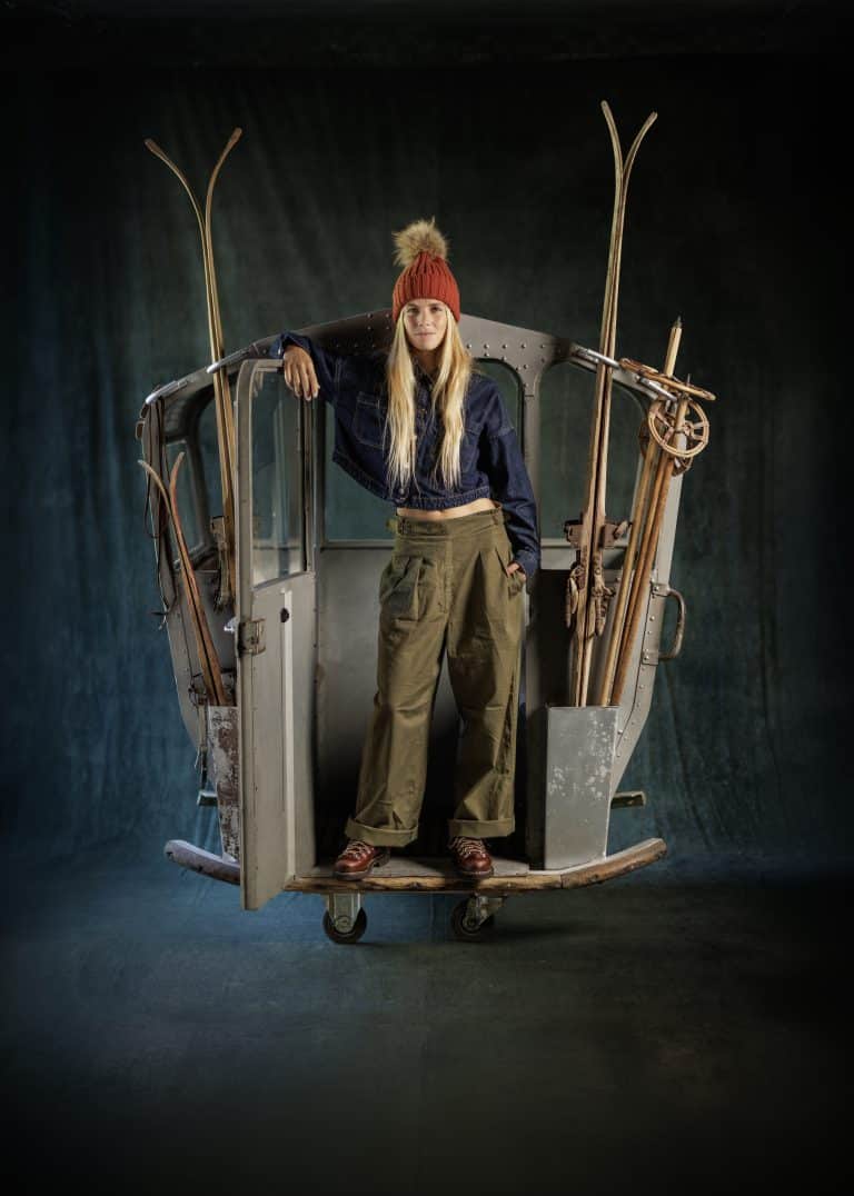Femme portant des clusaz Paraboot en cuir lisse écorce et une tenue d'inspiration vintage (bonnet rouge, veste denim, pantalon cargo vert) se tenant dans une ancienne télécabine de ski décorée de skis et bâtons en bois, sur un fond sombre.