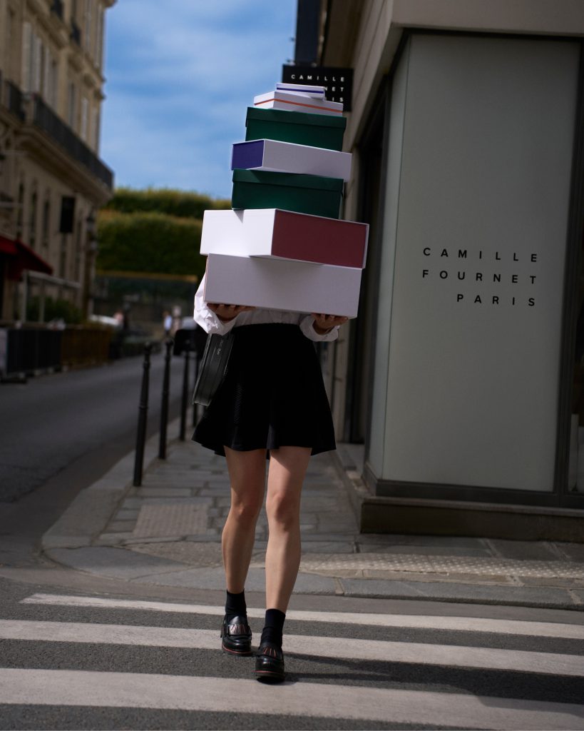 Femme traversant un passage piéton avec une pile de boîtes devant la boutique Camille Fournet Paris, portant les mocassins Orsay exclusifs.